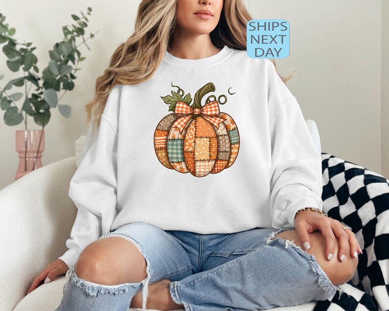 May include: White sweatshirt featuring a colorful patchwork pumpkin design with a bow and stem. The pumpkin is made of various patterned fabrics in orange, green, and brown. The text "SHIPS NEXT DAY" is visible.