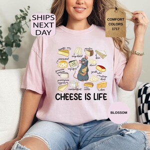 May include: A light pink t-shirt with a graphic of various cheeses and a cartoon rat holding cheese. The text "CHEESE IS LIFE" is below the graphic. The shirt also has the text "SHIPS NEXT DAY", "COMFORT COLORS 1717", and "BLOSSOM".