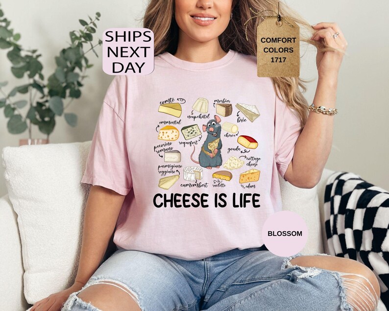 May include: A light pink t-shirt with a graphic of various cheeses and a cartoon rat holding cheese. The text "CHEESE IS LIFE" is below the graphic. The shirt also has the text "SHIPS NEXT DAY", "COMFORT COLORS 1717", and "BLOSSOM".