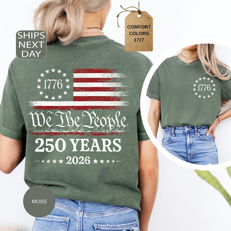 Pu&ograve; includere: T-shirt verde muschio con un disegno della bandiera americana. Il disegno include il testo "1776", "We The People" e "250 YEARS". La bandiera presenta strisce rosse e bianche e un cerchio di stelle. La maglietta ha un girocollo e maniche corte.