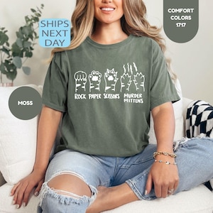 May include: A moss green t-shirt with a white graphic of rock, paper, scissors, and cat paws that say "Murder Mittens". The shirt is a comfort color and is paired with blue jeans. The text "Ships Next Day" is in a blue box.