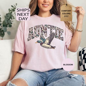 May include: A light pink t-shirt with the word "AUNTIE" in a camouflage pattern above a detailed illustration of a flying duck. The shirt has a relaxed fit and is made of a soft material. The text "SHIPS NEXT DAY" is in the upper left corner.