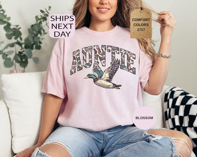 May include: A light pink t-shirt with the word "AUNTIE" in a camouflage pattern above a detailed illustration of a flying duck. The shirt has a relaxed fit and is made of a soft material. The text "SHIPS NEXT DAY" is in the upper left corner.