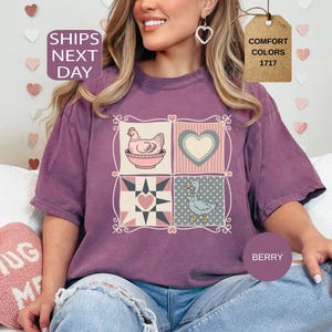 May include: Berry-coloured t-shirt with a patchwork design featuring a hen in a bowl, a heart, and a duck. The shirt has a relaxed fit and is made of a soft material. The text "Comfort Colors 1717" is on a tag.