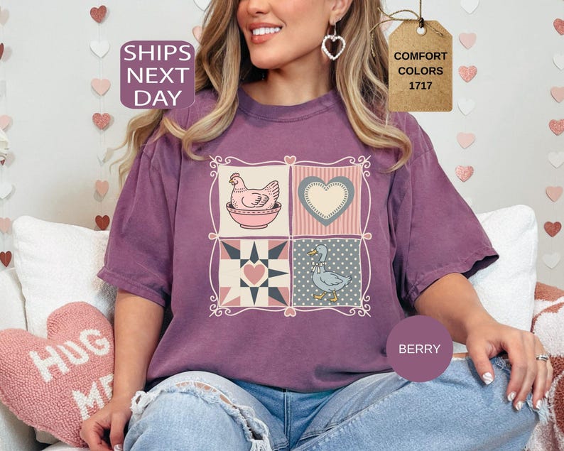May include: Berry-coloured t-shirt with a patchwork design featuring a hen in a bowl, a heart, and a duck. The shirt has a relaxed fit and is made of a soft material. The text "Comfort Colors 1717" is on a tag.