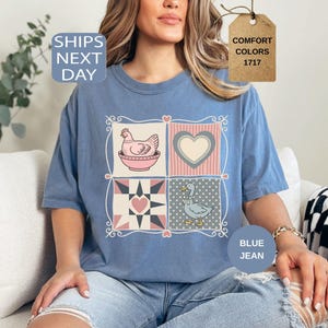 May include: Blue jean-coloured t-shirt with a patchwork design featuring a hen, a heart, a star, and a duck. The shirt has short sleeves and a crew neck. The text "Comfort Colors 1717" is on a tag.