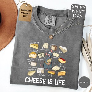 May include: A gray Comfort Colors t-shirt with a cheese-themed graphic and the text "CHEESE IS LIFE." The graphic features various cheese illustrations and a cartoon rat. A tag reads "COMFORT COLORS 1717."