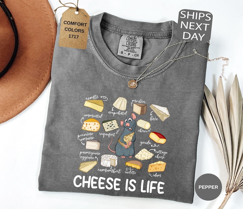 May include: A gray Comfort Colors t-shirt with a cheese-themed graphic and the text "CHEESE IS LIFE." The graphic features various cheese illustrations and a cartoon rat. A tag reads "COMFORT COLORS 1717."
