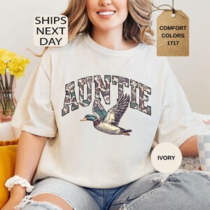 May include: Ivory-colored t-shirt with the word "AUNTIE" in a camouflage-style font, above a detailed illustration of a flying duck. The shirt also features the text "COMFORT COLORS 1717" on a tag. The words "SHIPS NEXT DAY" are also visible.