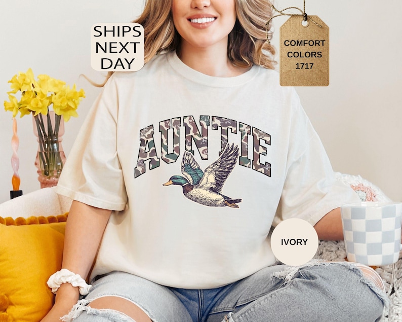May include: Ivory-colored t-shirt with the word "AUNTIE" in a camouflage-style font, above a detailed illustration of a flying duck. The shirt also features the text "COMFORT COLORS 1717" on a tag. The words "SHIPS NEXT DAY" are also visible.