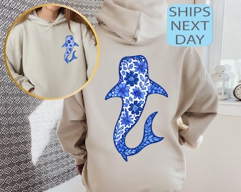 May include: Beige hoodie featuring a blue floral whale shark design on the back and a smaller version on the front. The whale shark design is filled with blue flowers and leaves. The hoodie has a drawstring hood and a kangaroo pocket.
