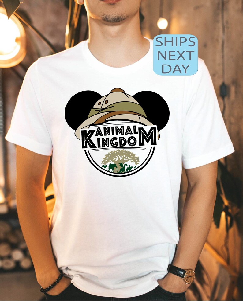 May include: White t-shirt featuring a black and green Animal Kingdom logo with Mickey Mouse ears and a safari hat. The logo includes a tree with animals surrounding it.