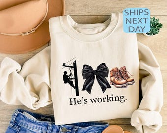 May include: Cream-coloured sweatshirt featuring a lineman graphic on a utility pole, a black bow, and work boots. The text "He's working" is printed below the graphic. The sweatshirt is displayed on a wooden surface.