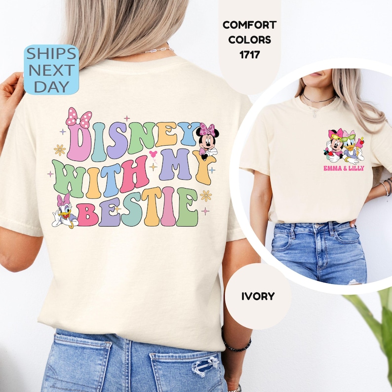 May include: Ivory t-shirt with the text "DISNEY WITH MY BESTIE" in rainbow colors. The back of the shirt features a small image of Minnie Mouse and Daisy Duck. The front of the shirt features a small image of Minnie Mouse and Daisy Duck with the text "Emma & Lilly".