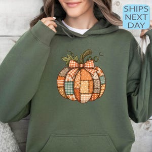 May include: An olive green hoodie featuring a patchwork pumpkin design with a plaid bow. The pumpkin has orange, brown, and green patches. The text "SHIPS NEXT DAY" is in a blue box.