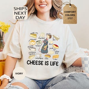May include: Ivory-colored t-shirt featuring a cartoon rat holding cheese, surrounded by illustrations of various cheeses. The text "CHEESE IS LIFE" is printed below the rat. The shirt also has the text "COMFORT COLORS 1717".