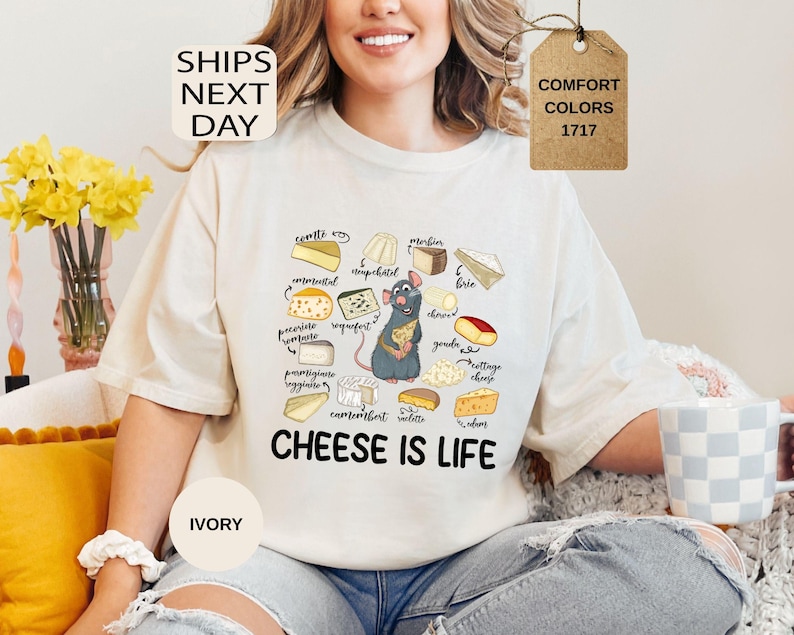 May include: Ivory-colored t-shirt featuring a cartoon rat holding cheese, surrounded by illustrations of various cheeses. The text "CHEESE IS LIFE" is printed below the rat. The shirt also has the text "COMFORT COLORS 1717".