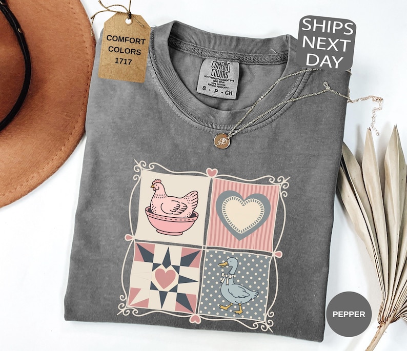 May include: A grey Comfort Colors t-shirt with a patchwork design featuring a chicken in a bowl, a heart, and a duck. The shirt has a crew neck and short sleeves. A tag reads "Comfort Colors 1717". A necklace is draped across the shirt.