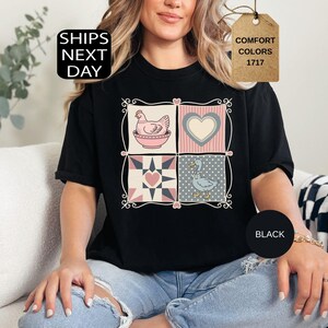 May include: Black t-shirt with a four-panel design: a hen in a bowl, a heart, a quilt pattern, and a goose. The shirt has the text "Comfort Colors 1717" on a tag and "Ships Next Day" on a black square.