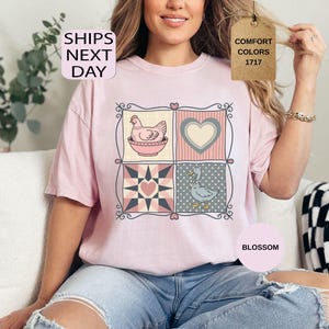 May include: A pale pink t-shirt with a patchwork design featuring a hen, a heart, a star, and a goose. The shirt has the text "SHIPS NEXT DAY" and "COMFORT COLORS 1717". The word "BLOSSOM" is also visible.