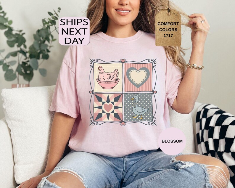 May include: A pale pink t-shirt with a patchwork design featuring a hen, a heart, a star, and a goose. The shirt has the text "SHIPS NEXT DAY" and "COMFORT COLORS 1717". The word "BLOSSOM" is also visible.