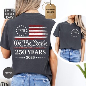 Pu&ograve; includere: T-shirt grigio scuro con grafica della bandiera americana, testo "1776", "We The People" e "250 YEARS 2026". La maglietta ha girocollo e maniche corte. Il colore &egrave; "Pepper".