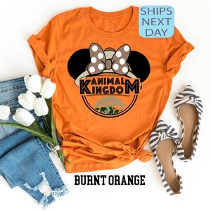 May include: Burnt orange t-shirt featuring a white and black polka dot Minnie Mouse ears design and the text "Animal Kingdom" in a black circle with a green tree and animals inside.