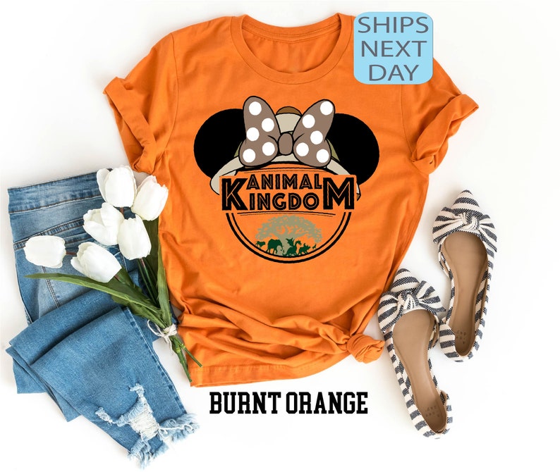 May include: Burnt orange t-shirt featuring a white and black polka dot Minnie Mouse ears design and the text "Animal Kingdom" in a black circle with a green tree and animals inside.