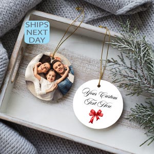 May include: Two round Christmas ornaments. One features a family photo, the other has the text "Your Custom Text Here" with a red bow. Both have gold string hangers. The ornaments rest on a white tray.