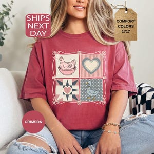 May include: Crimson t-shirt with a patchwork design featuring a hen, heart, and goose. The shirt has the text "SHIPS NEXT DAY" and "COMFORT COLORS 1717" on a tag. The shirt is a relaxed fit.