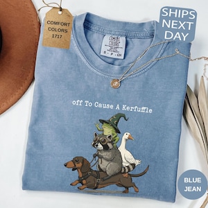 Può includere: Una t-shirt color blu jeans con una grafica raffigurante un bassotto, un procione con occhiali protettivi e una rana con un cappello da mago sulla schiena del cane, insieme a un'oca bianca. Il testo riporta off To Cause A Kerfuffle.