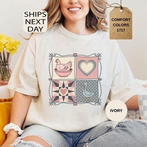 May include: Ivory t-shirt with a patchwork design featuring a hen, heart, and duck. The shirt has the text "SHIPS NEXT DAY" and "COMFORT COLORS 1717". The t-shirt is a comfortable fit.