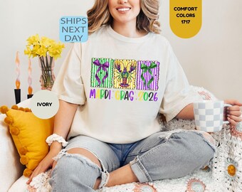 May include: An ivory short-sleeve t-shirt featuring a graphic print of three rectangular frames with purple and green stripes. The center frame contains a crawfish, while the outer frames feature bows. The text reads MARDI GRAS 2026.