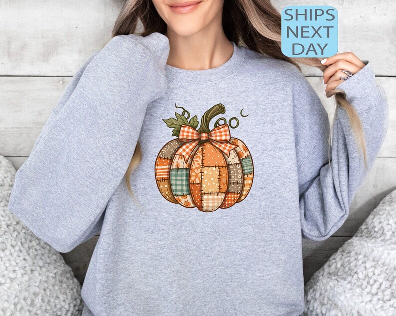 May include: A light gray sweatshirt featuring a colorful pumpkin design. The pumpkin is made of patchwork fabric in shades of orange, brown, and green, with a plaid bow and stem. The text "SHIPS NEXT DAY" is in a blue box.