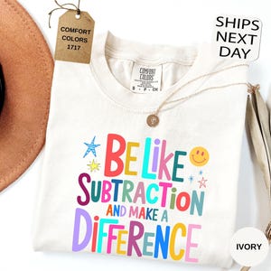 May include: An ivory t-shirt featuring the multi-colored text "BE LIKE SUBTRACTION AND MAKE A DIFFERENCE" in various fonts. The design includes small star and smiley face graphics. A brown tag labeled "COMFORT COLORS 1717" is attached to the collar.