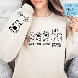 May include: Cream-colored sweatshirt with black paw print graphics. The left sleeve has a paw print and the name "Brady" in script. The front features paw prints with the text "Rock Paper Scissors Murder Mittens".