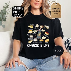 May include: Black t-shirt with a graphic of various cheeses and a cartoon rat holding a cheese wedge. The text "CHEESE IS LIFE" is below the graphic. The shirt also has the text "SHIPS NEXT DAY" and "COMFORT COLORS 1717".