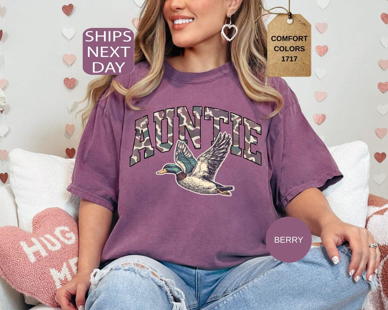 May include: Berry-colored Auntie t-shirt with a duck graphic. The word "AUNTIE" is printed in a camouflage font. The shirt has a relaxed fit and is made of a soft material. The image also includes the text "SHIPS NEXT DAY" and "COMFORT COLORS 1717".