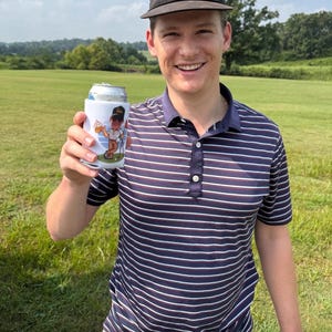 May include: A white foam beverage insulator featuring a graphic of a cartoon golfer holding a beer and a golf club. The insulator is held up, displaying the colourful illustration against a dark blue, white, and red striped polo shirt.
