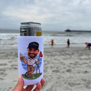 May include: A white foam beverage can cooler featuring a cartoon illustration of a bearded person wearing a dark cap, a white polo shirt, and brown trousers, holding a beer mug next to a golf flag on a green patch against a blue circular background.