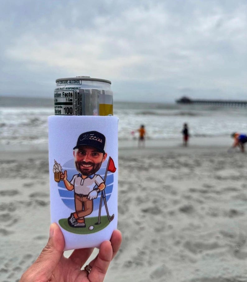 May include: A white foam beverage can cooler featuring a cartoon illustration of a bearded person wearing a dark cap, a white polo shirt, and brown trousers, holding a beer mug next to a golf flag on a green patch against a blue circular background.