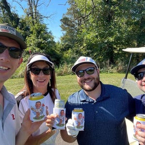 May include: Four people wearing sunglasses and hats hold white beverage can coolers featuring a cartoon illustration of a golfer. The coolers are held in front of the individuals, who are positioned outdoors in a grassy area near a golf cart.