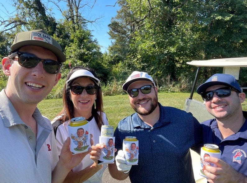 May include: Four people wearing sunglasses and hats hold white beverage can coolers featuring a cartoon illustration of a golfer. The coolers are held in front of the individuals, who are positioned outdoors in a grassy area near a golf cart.