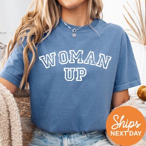 A woman wearing a blue t-shirt with the text "WOMAN UP" printed on it.