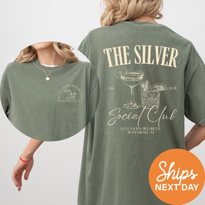 May include: An olive green t-shirt featuring a cream-colored graphic on the back with the text THE SILVER Social Club, EST 2024, and KAYLA'S BACHELORETTE SCOTTSDALE AZ, alongside illustrations of a martini glass and a rocks glass with a lime wedge.