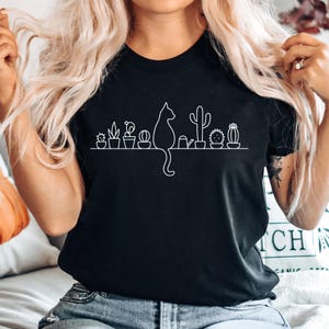 A woman with long blonde hair is wearing a black t-shirt with a graphic design of a cat and various cacti and succulents.