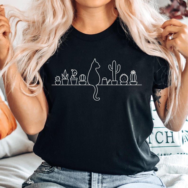 A woman with long blonde hair is wearing a black t-shirt with a graphic design of a cat and various cacti and succulents.