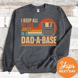May include: Dark gray sweatshirt with the text "I KEEP ALL MY DAD JOKES IN A DAD-A-BASE" in a retro design. The design includes a floppy disk graphic and a sunset-style color scheme. The sweatshirt is a crew neck style.