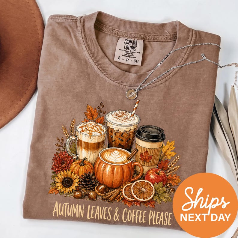 A t-shirt with a graphic design depicting autumn leaves and coffee, along with a necklace and a hat, all set against a white background.