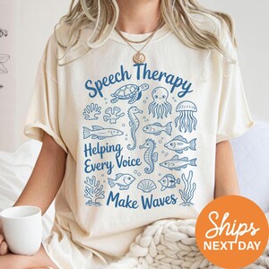 A person wearing a white t-shirt with a graphic design featuring various sea creatures and the text "Speech Therapy Helping Every Voice Make Waves" printed on it.